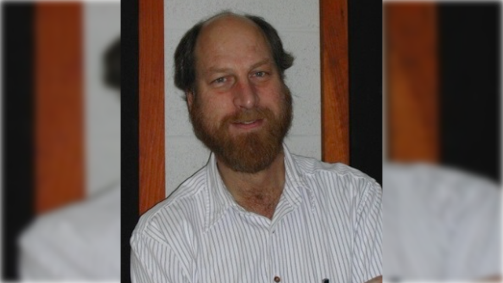 Portrait of a man with a beard, wearing a striped shirt, smiling against a backdrop of wooden and gray walls.