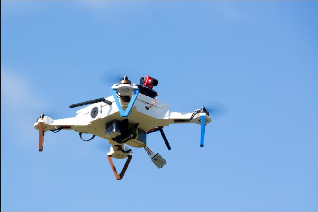 A white drone equipped with a camera and sensors flying against a clear blue sky.
