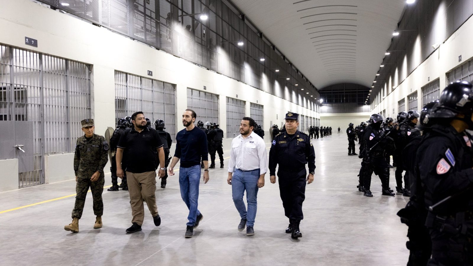 Security officials and military personnel walk through a high-security prison facility, observing inmates and law enforcement officers lined up in the background.