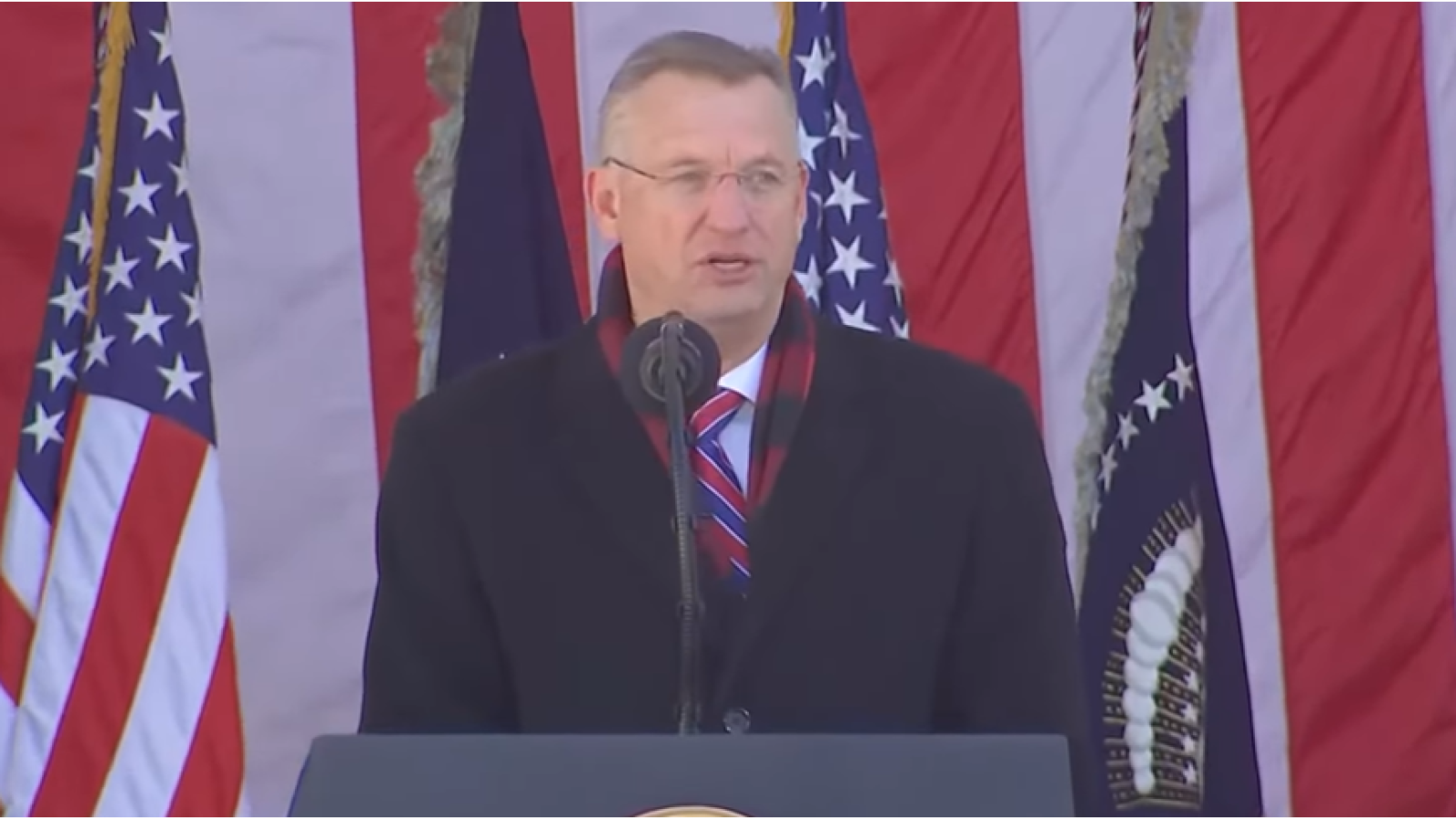 Man in a black coat and glasses delivering a speech at an outdoor event, with American flags and a presidential seal in the background.