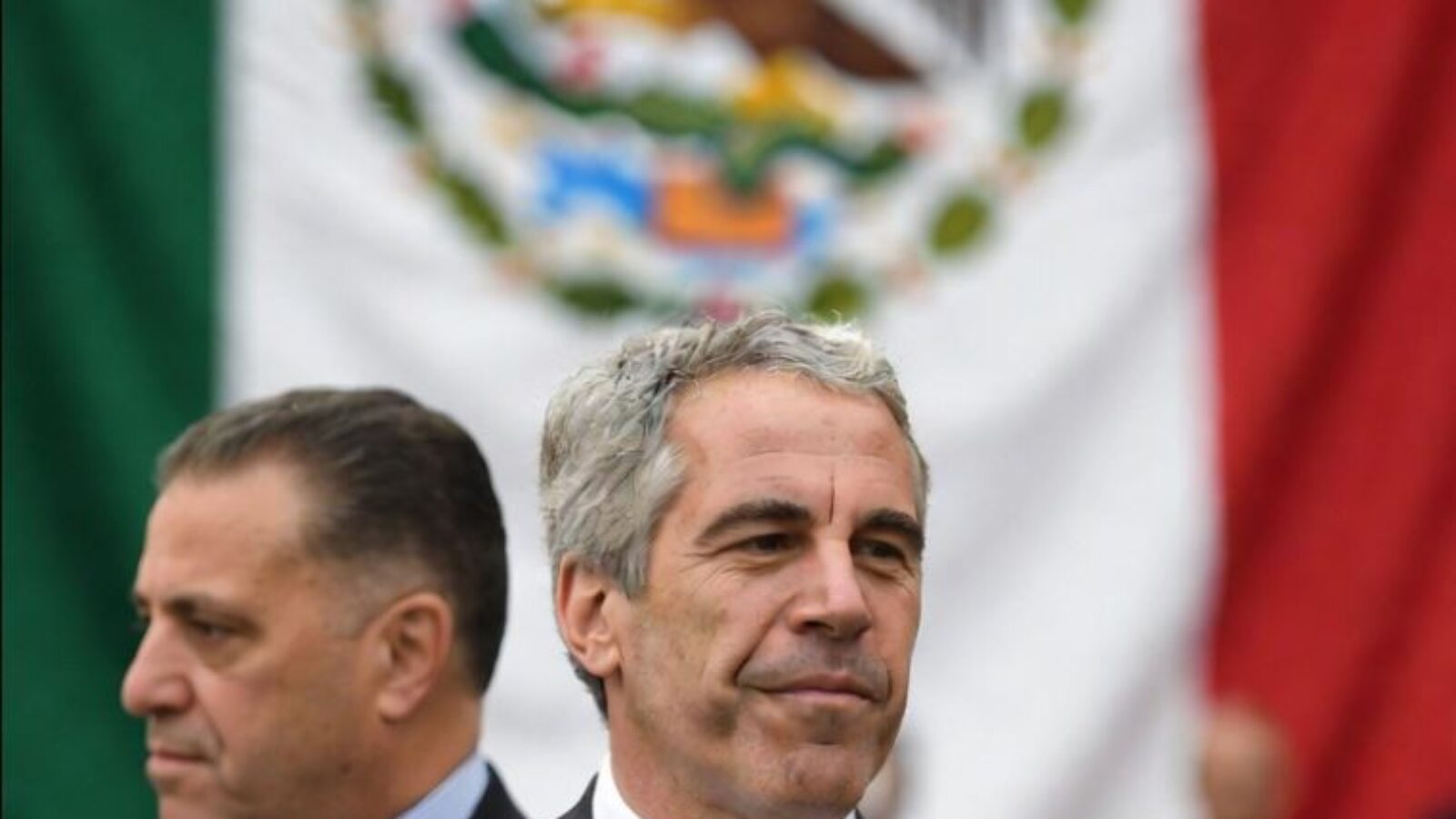 Two men in formal attire stand in front of the Mexican flag, emphasizing a cultural or political event.