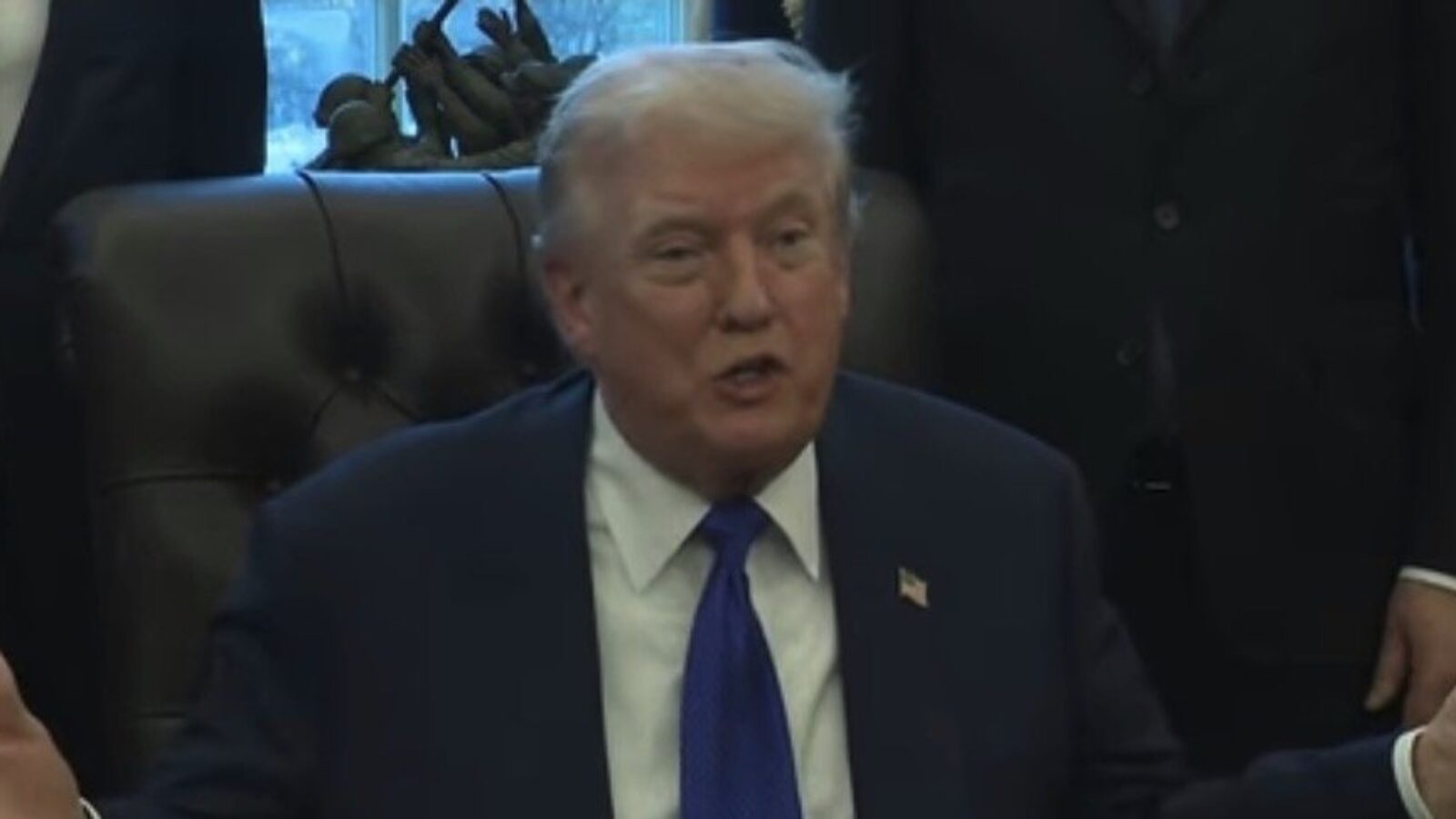 Donald Trump speaking during a meeting in the Oval Office, wearing a suit and tie, with attendees in the background.