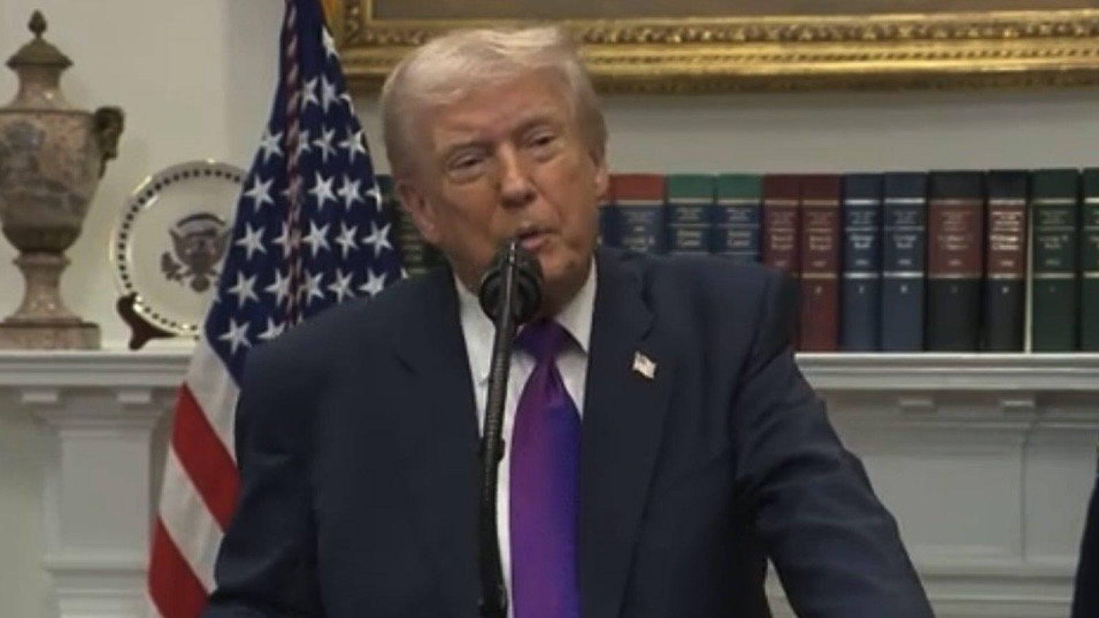 Donald Trump speaking at a podium with the American flag in the background, addressing an audience in a formal setting.