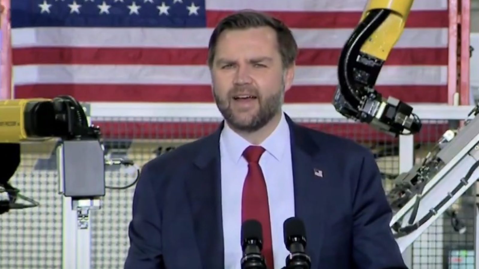 A speaker addresses an audience at a manufacturing facility, with robotic arms and an American flag in the background, highlighting advancements in technology and industry.