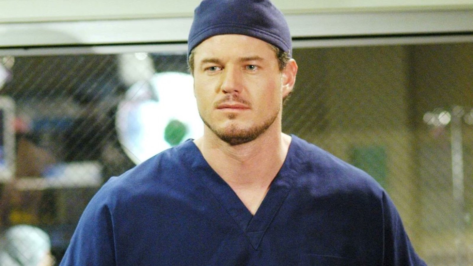 Male surgeon in blue scrubs and cap, looking serious in a hospital setting, with medical equipment blurred in the background.