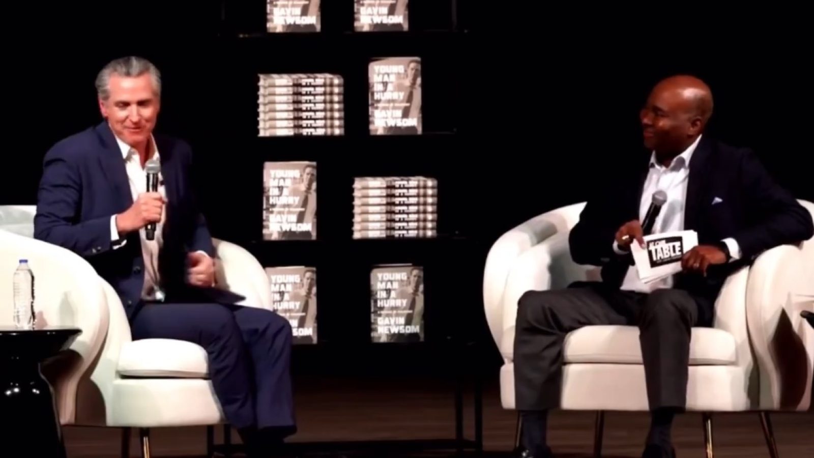 Gavin Newsom participates in a discussion at an event promoting his book, seated alongside another speaker in front of a display of books.
