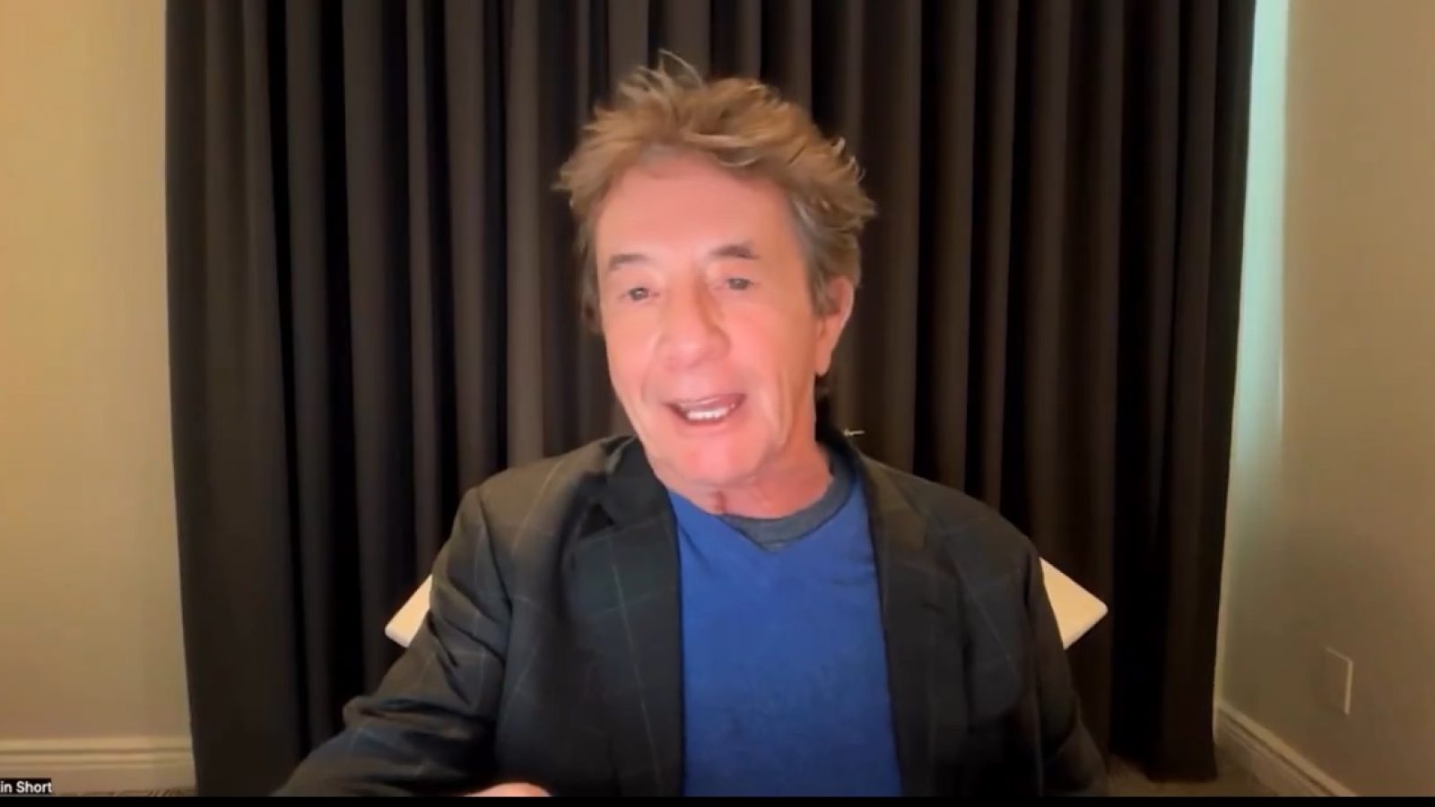 Martin Short speaks during a virtual event, wearing a blazer over a blue shirt, with dark curtains in the background.