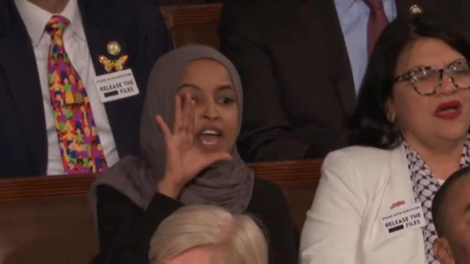 Ilhan Omar gestures passionately while speaking during a congressional event, with other attendees visible in the background.