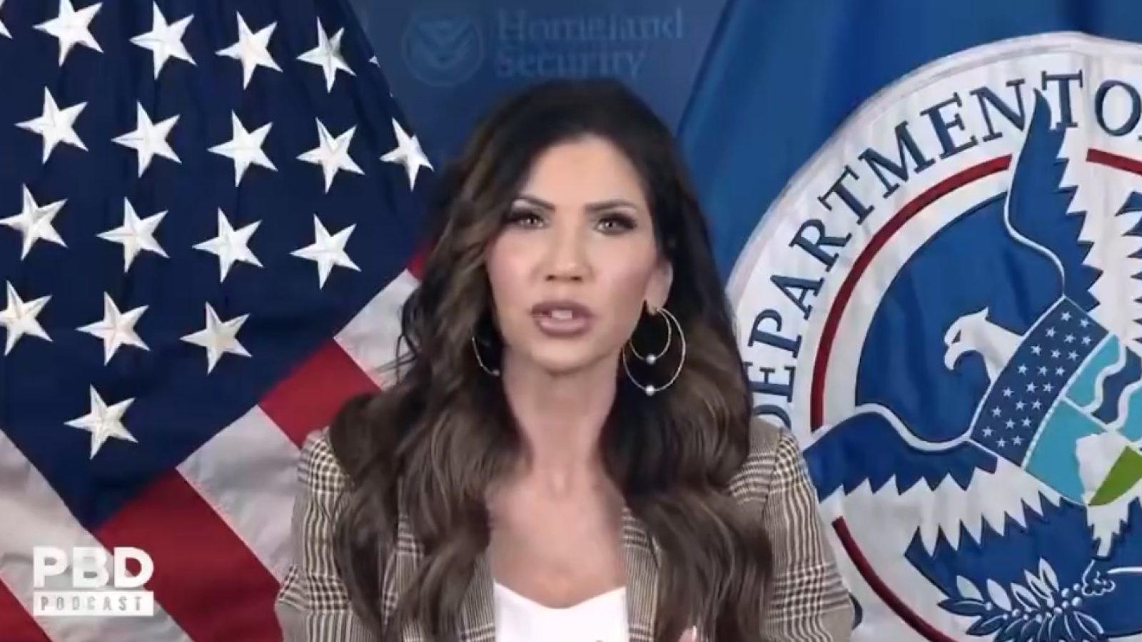 Woman speaking at a podium with the Department of Homeland Security seal and an American flag backdrop during a podcast discussion.