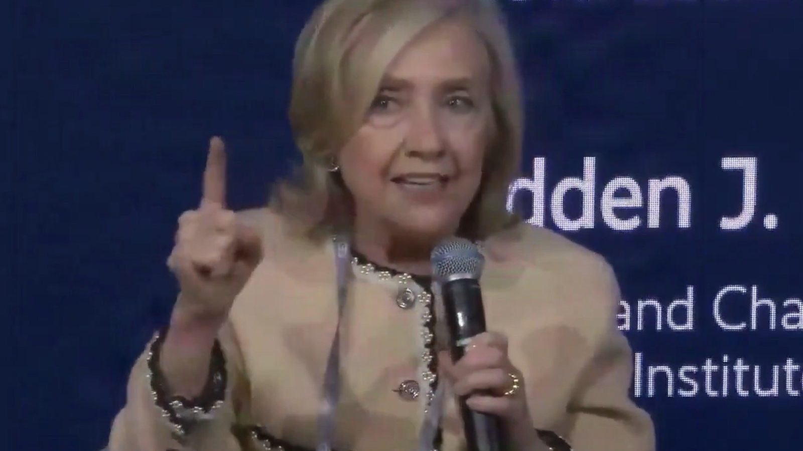Hillary Clinton speaking passionately at a conference, holding a microphone and gesturing with one hand to emphasize her point.