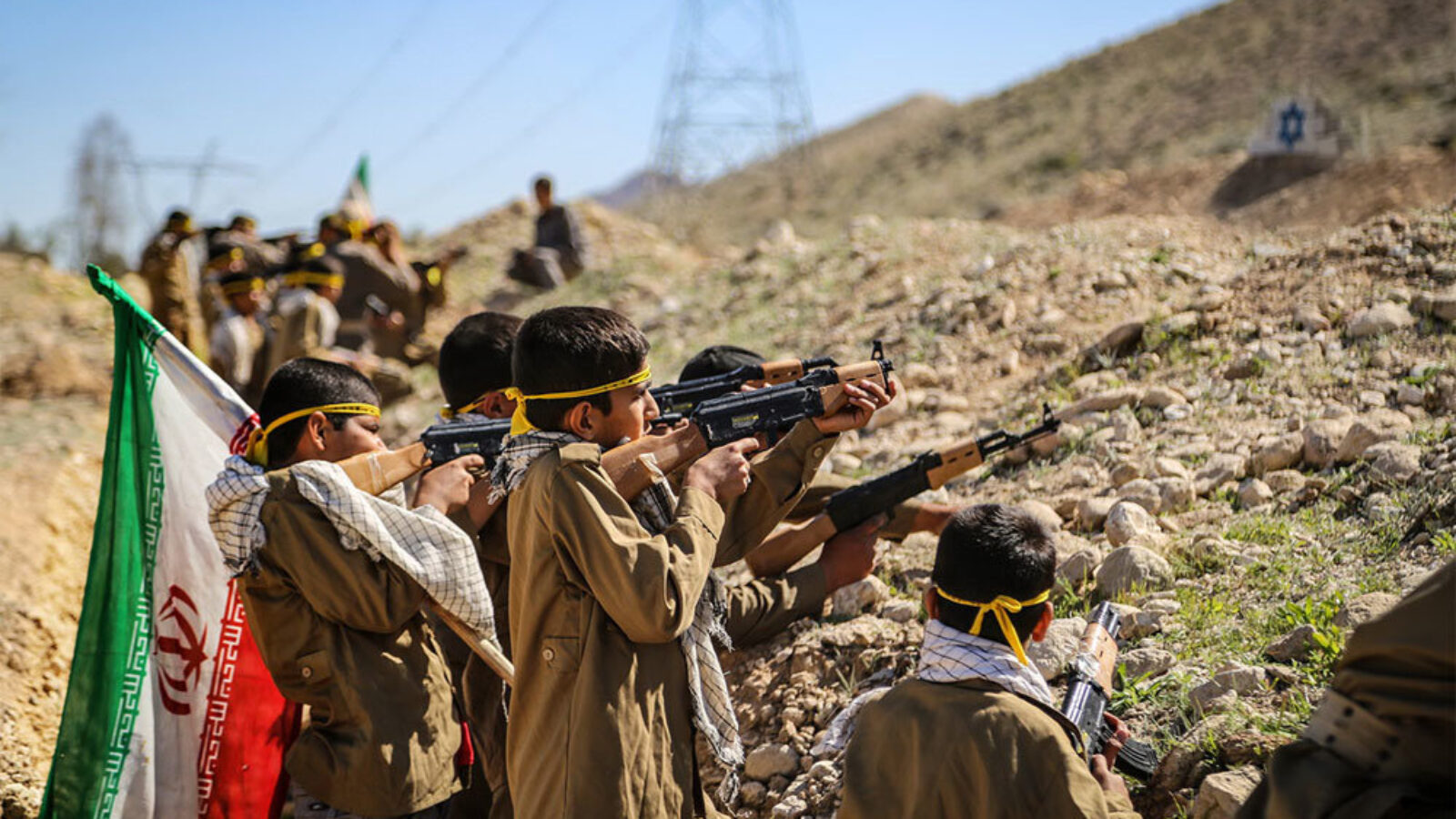 Children in military attire practice shooting with rifles during a training exercise, with flags in the background.