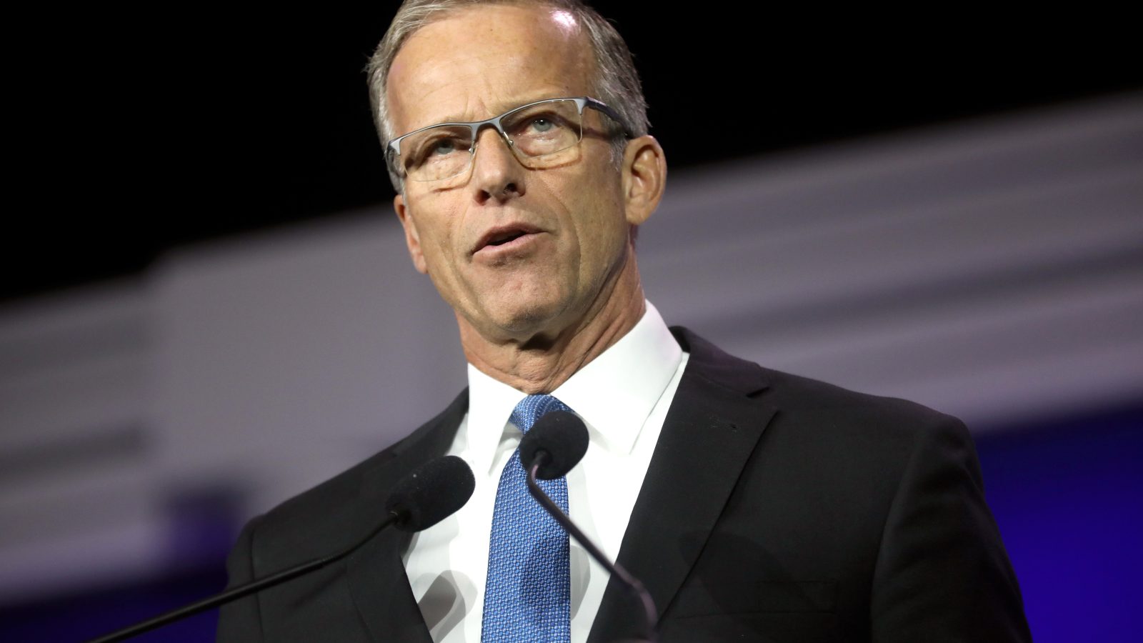 Business professional delivering a speech at a conference, wearing glasses and a suit, with microphones in front of him.