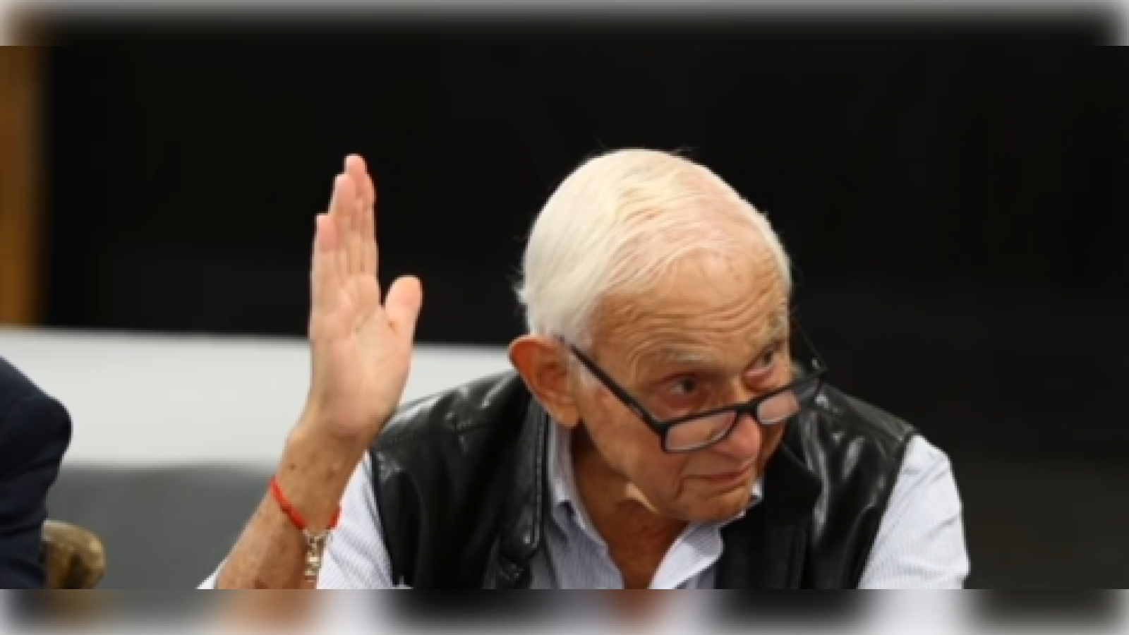Elderly man with glasses raising his hand during a discussion, wearing a black vest and a light blue shirt, in a meeting setting.