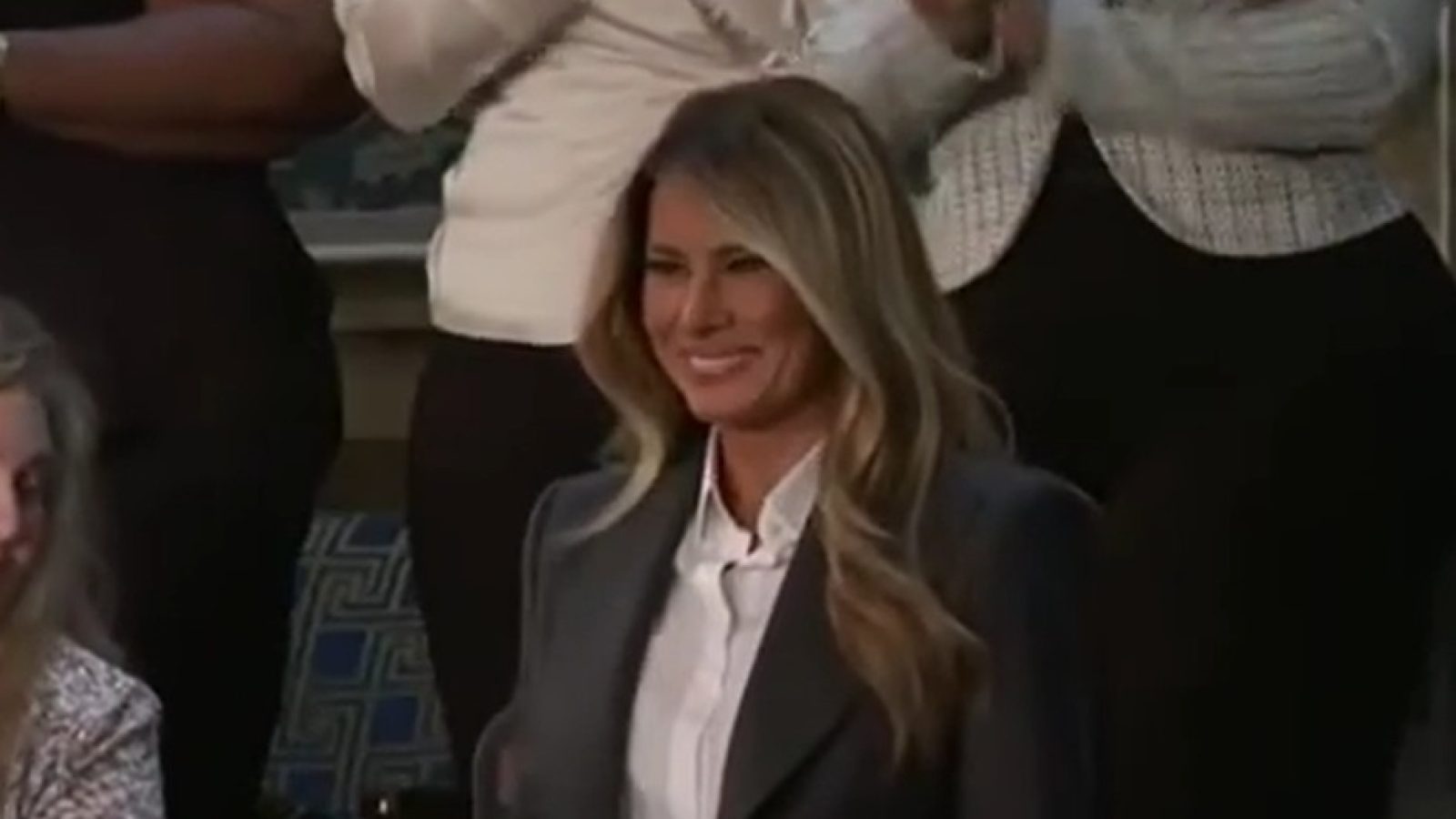 Melania Trump smiling while attending an event, surrounded by applauding audience members, wearing a stylish outfit with long hair.