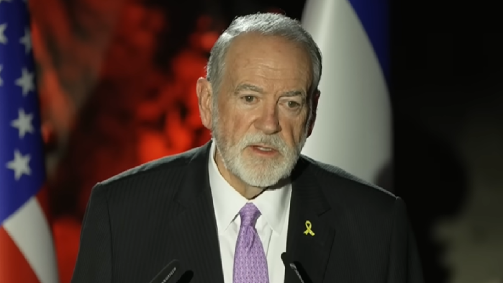 A man in formal attire with a beard speaks at a podium, featuring the American flag and a yellow ribbon, during an event.