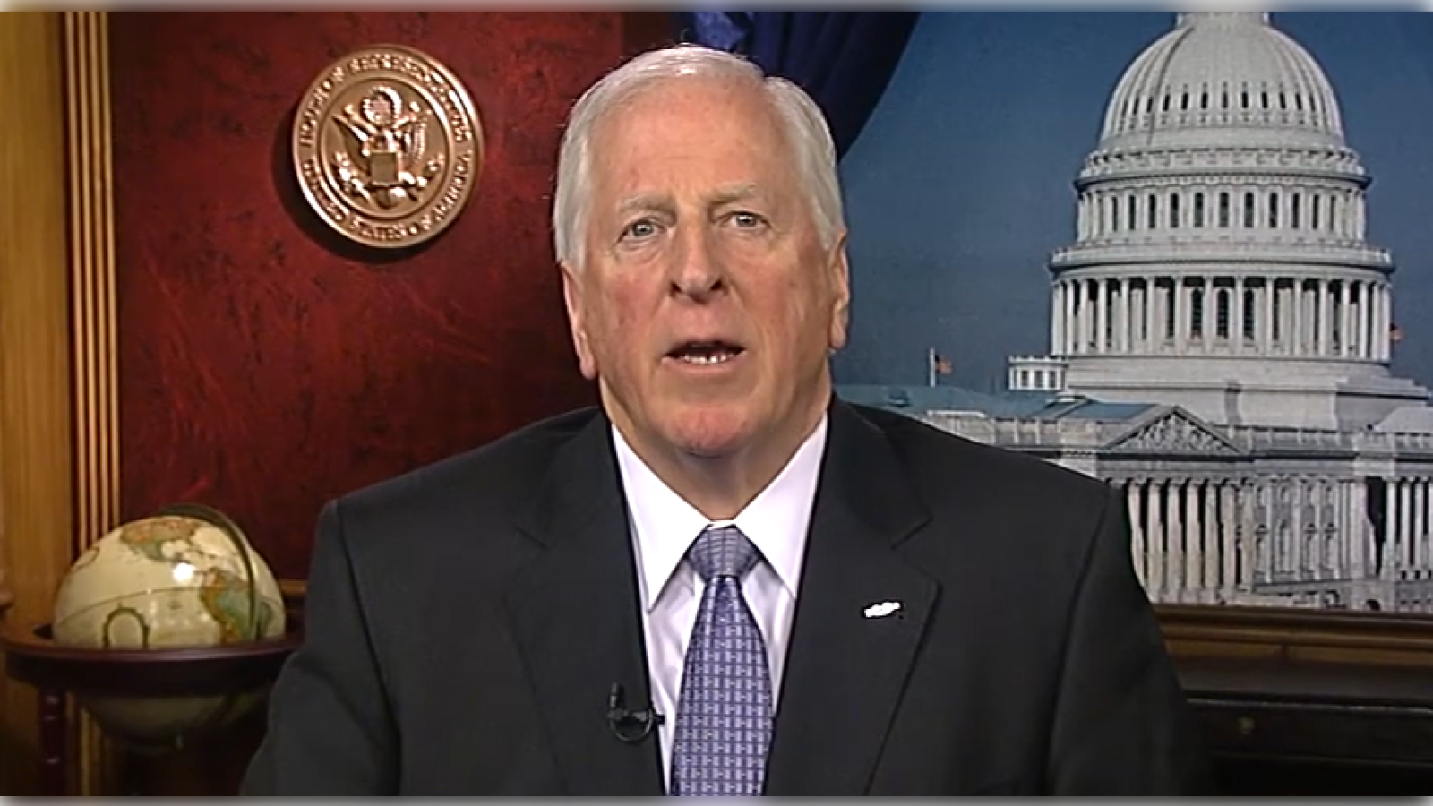 Man in a suit speaking from an office with a view of the U.S. Capitol, showcasing a globe and official seal in the background.