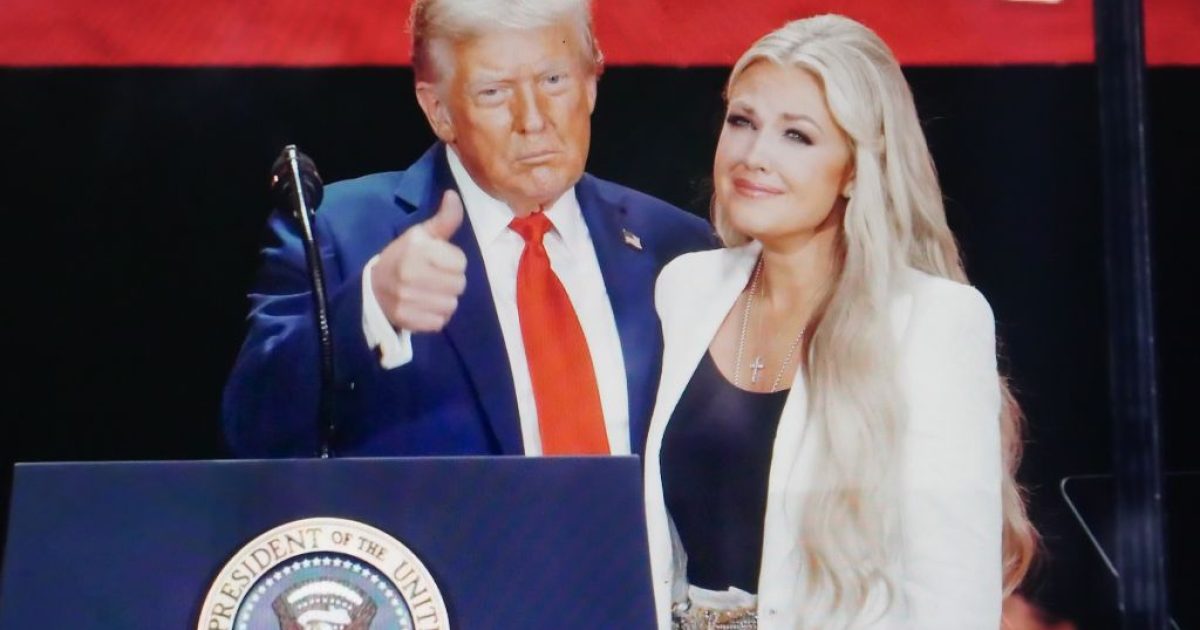 Donald Trump giving a thumbs up while standing next to a woman in a white blazer at a public speaking event with a presidential seal backdrop.