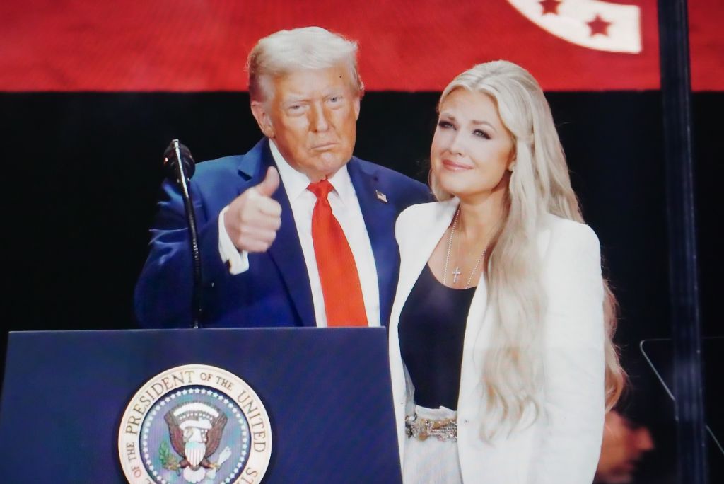 Donald Trump giving a thumbs up while standing next to a woman in a white blazer at a public speaking event with a presidential seal backdrop.