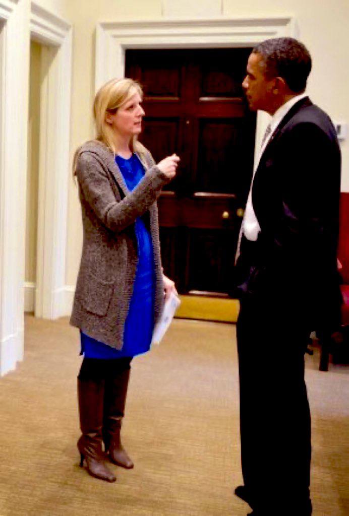 Woman in a blue dress and gray cardigan converses with a man in a suit inside an office setting.