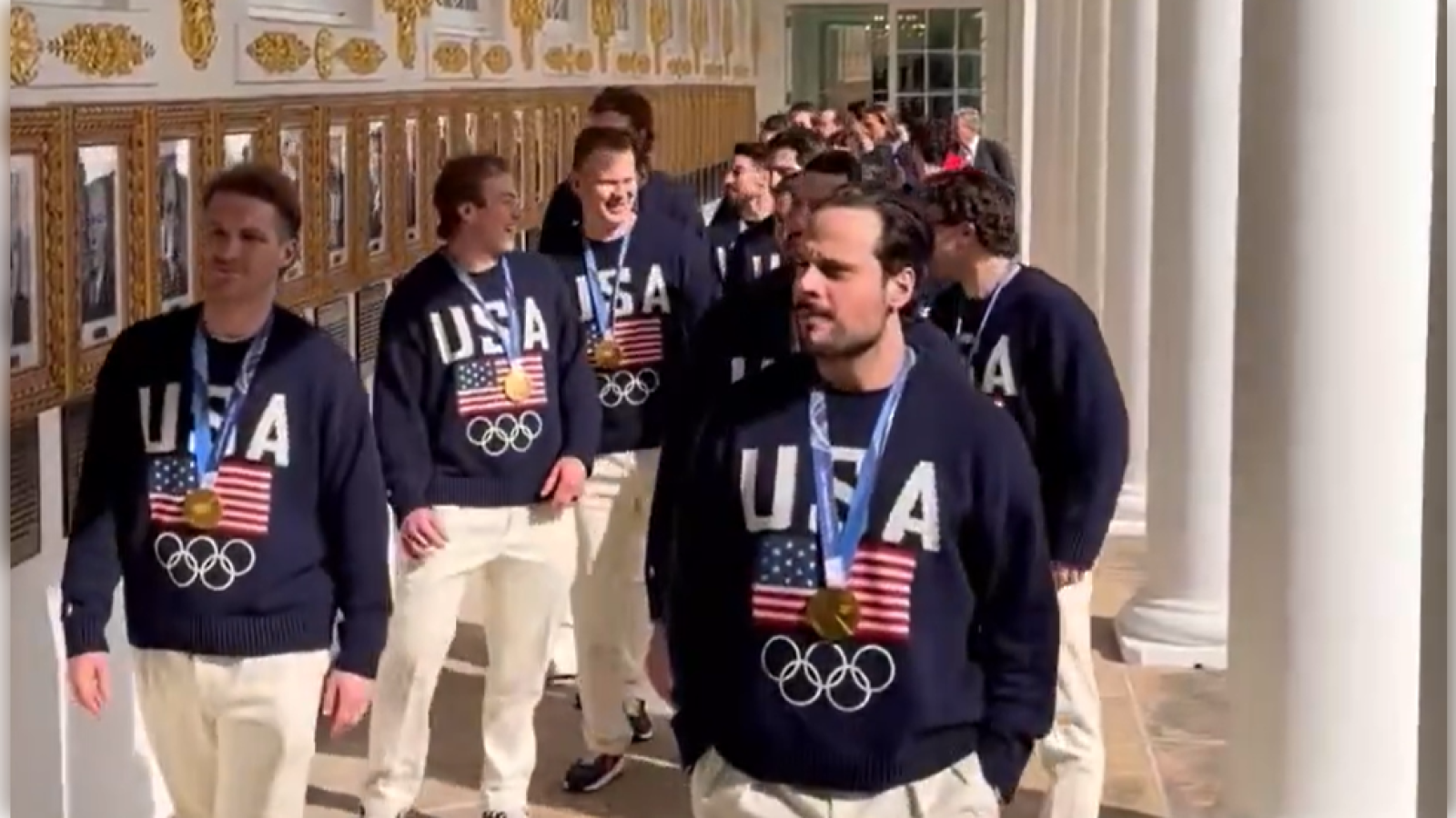 U.S. Olympic athletes in navy sweaters with gold medals walk past historical photos in a grand hall, showcasing their achievements and national pride.