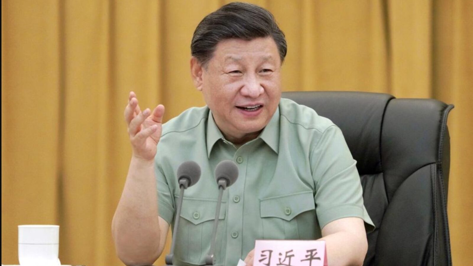 Chinese President Xi Jinping speaking during a meeting, gesturing with his hand, seated at a table with a cup and documents, against a backdrop of golden curtains.