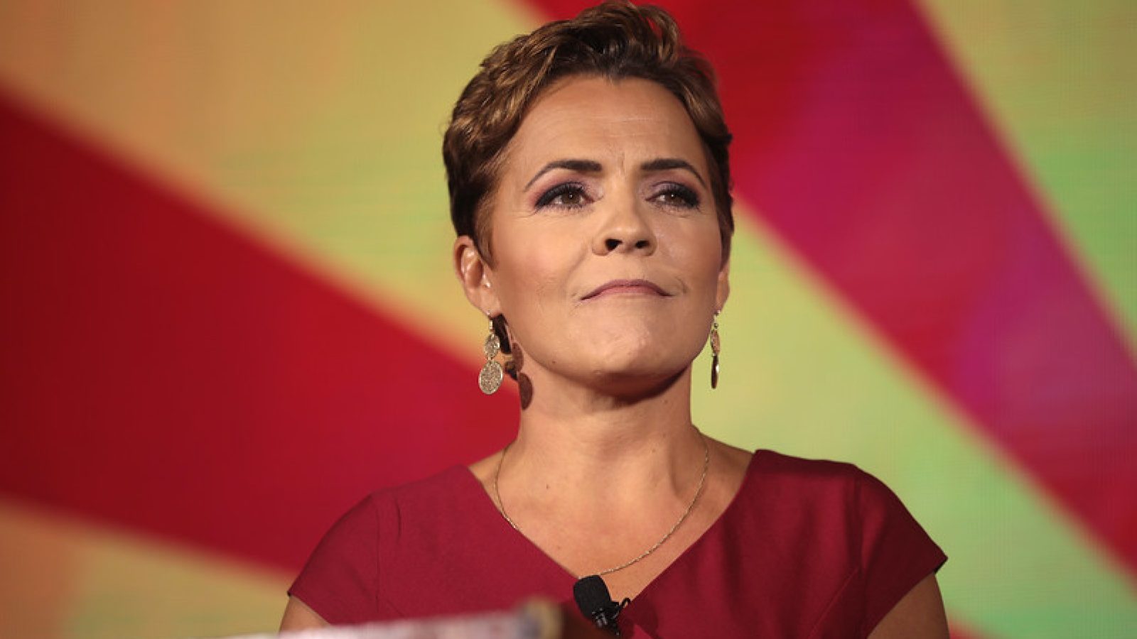 A confident woman in a red dress stands at a podium, delivering a speech against a vibrant background featuring rays of color.