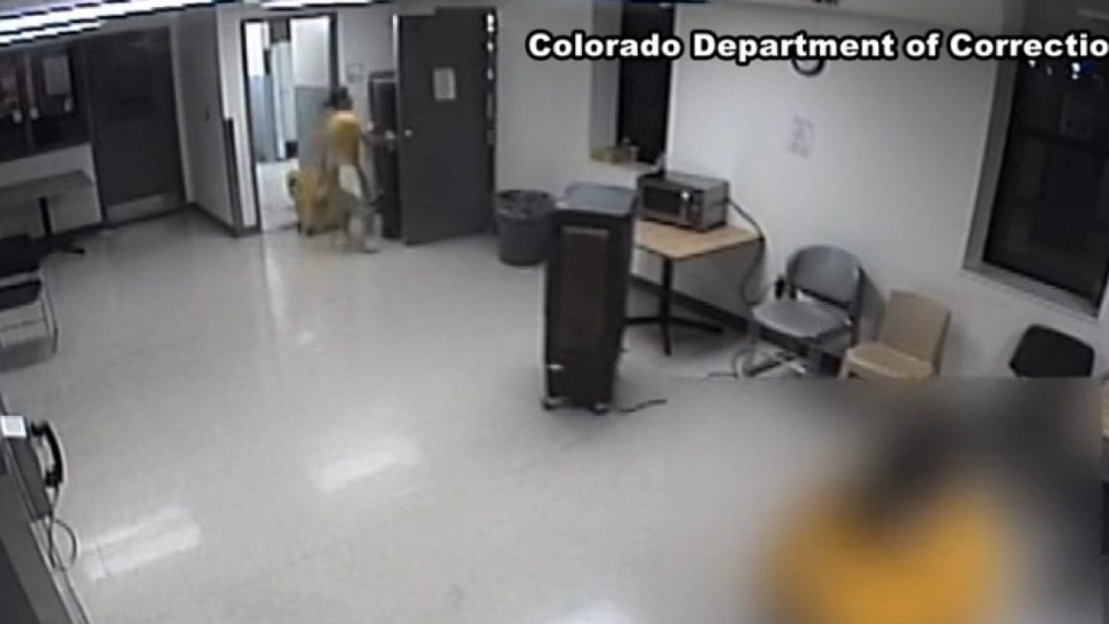 Surveillance footage shows a hallway in a correctional facility with staff and inmates, highlighting the interior layout and security measures in place.