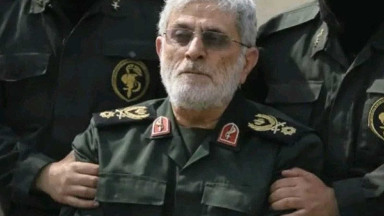 Iranian military officer in uniform with insignia, appearing serious while being assisted by two others in military attire.