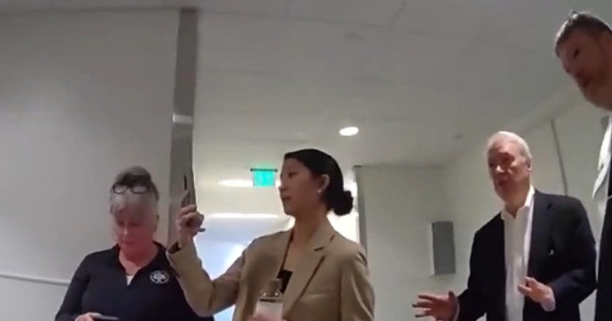 Group of four individuals engaged in conversation in a hallway, with one person recording on a smartphone and others discussing.