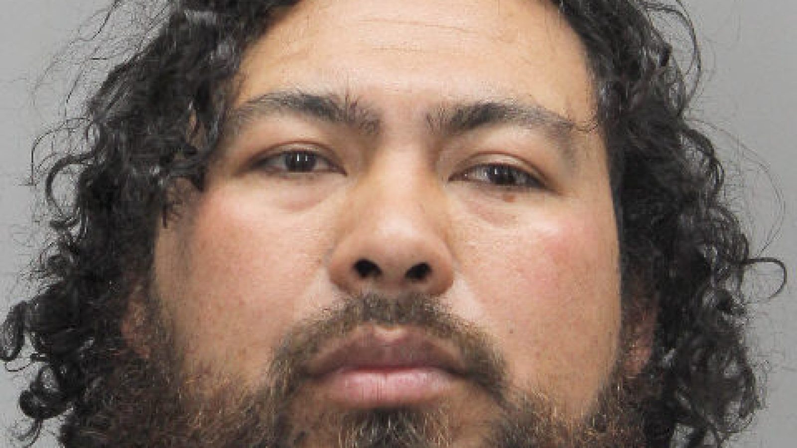 Mugshot of a man with curly hair and a beard, wearing a white garment against a gray background.