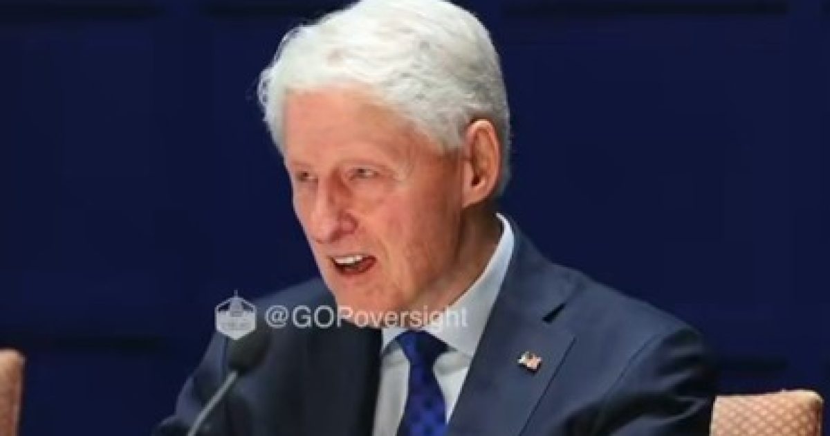 Bill Clinton speaking at a formal event, wearing a dark suit with a blue tie, highlighting his engagement in discussion.