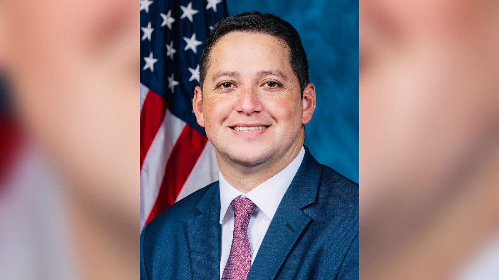 Portrait of a smiling man in a suit with a pink tie, standing in front of an American flag backdrop, representing a professional political figure.