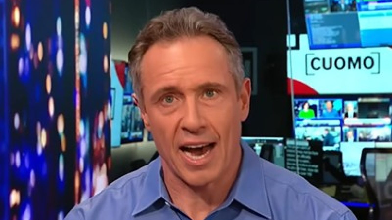 News anchor Chris Cuomo delivers a report in a television studio with multiple screens displaying news content in the background.