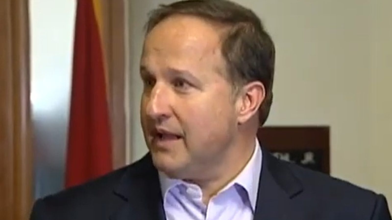 Man speaking during a press conference, wearing a dark blazer and light shirt, with flags visible in the background.