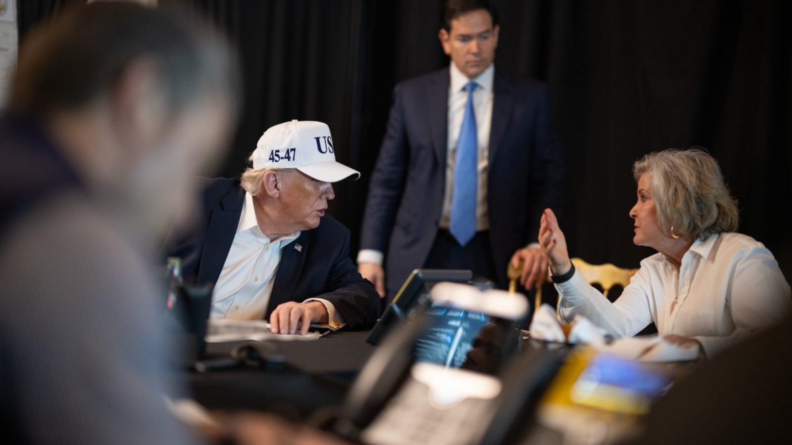 Donald Trump, wearing a white cap, engages in conversation with a woman at a table while another man observes, surrounded by phones and technology.