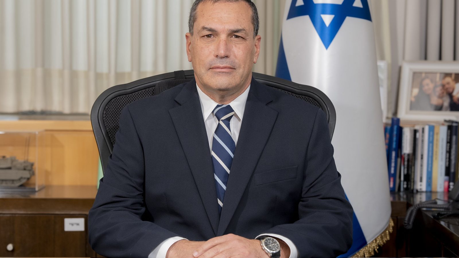 Portrait of a man in a suit sitting at a desk, with an Israeli flag in the background, conveying leadership and professionalism.