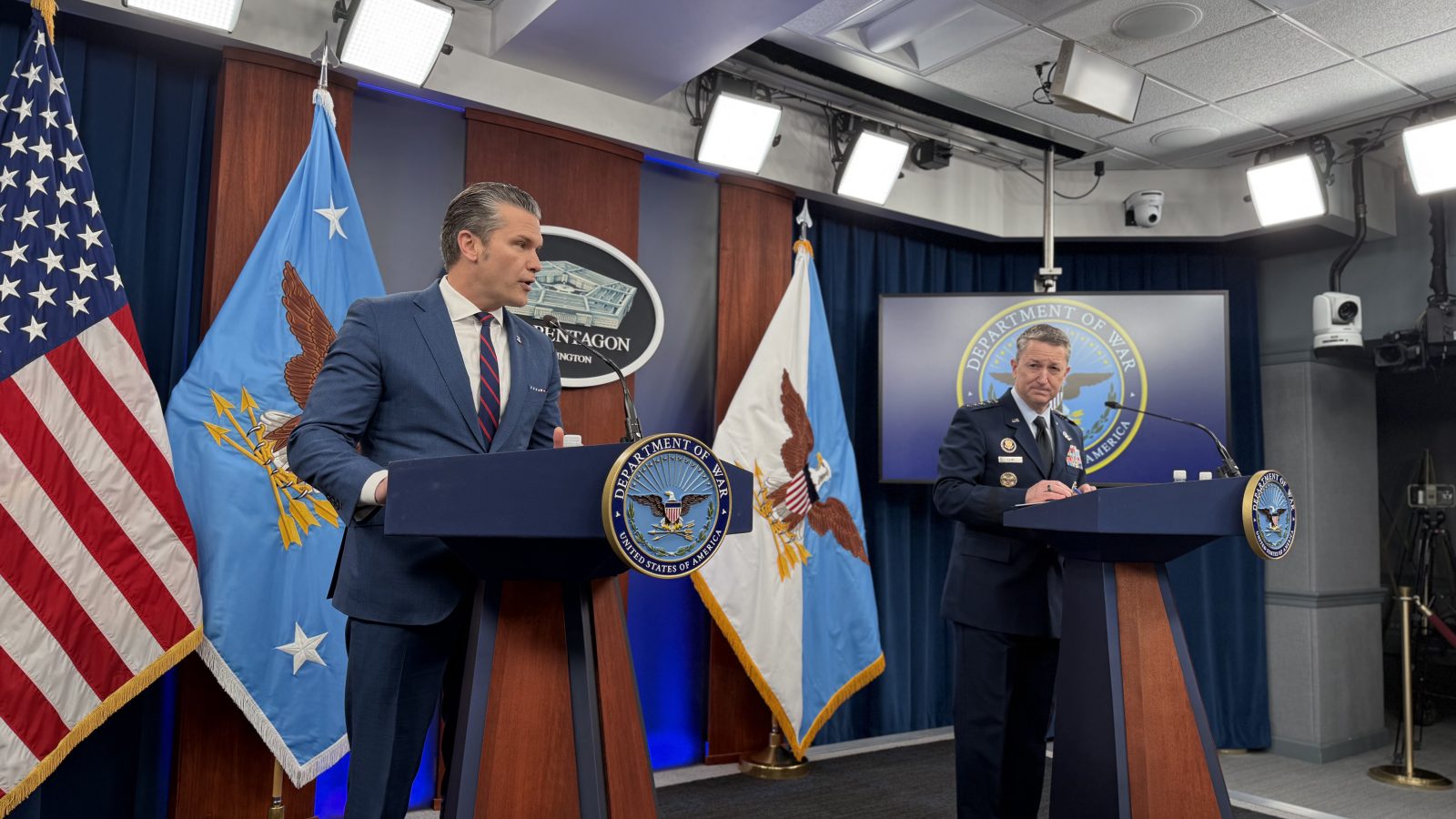 Press conference at the Pentagon featuring two officials, one in a suit and the other in military uniform, with American and Air Force flags in the background.