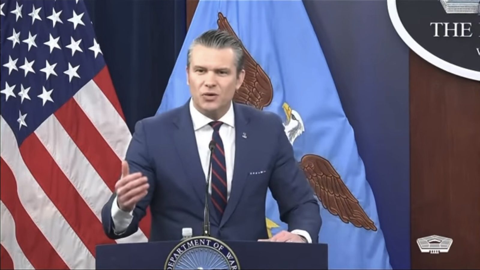 U.S. Department of Defense spokesperson delivering a statement at a press briefing, with American and Air Force flags in the background.