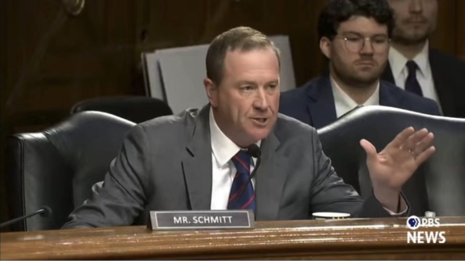 Senator Schmitt speaks passionately during a congressional hearing, with a colleague attentively listening in the background.