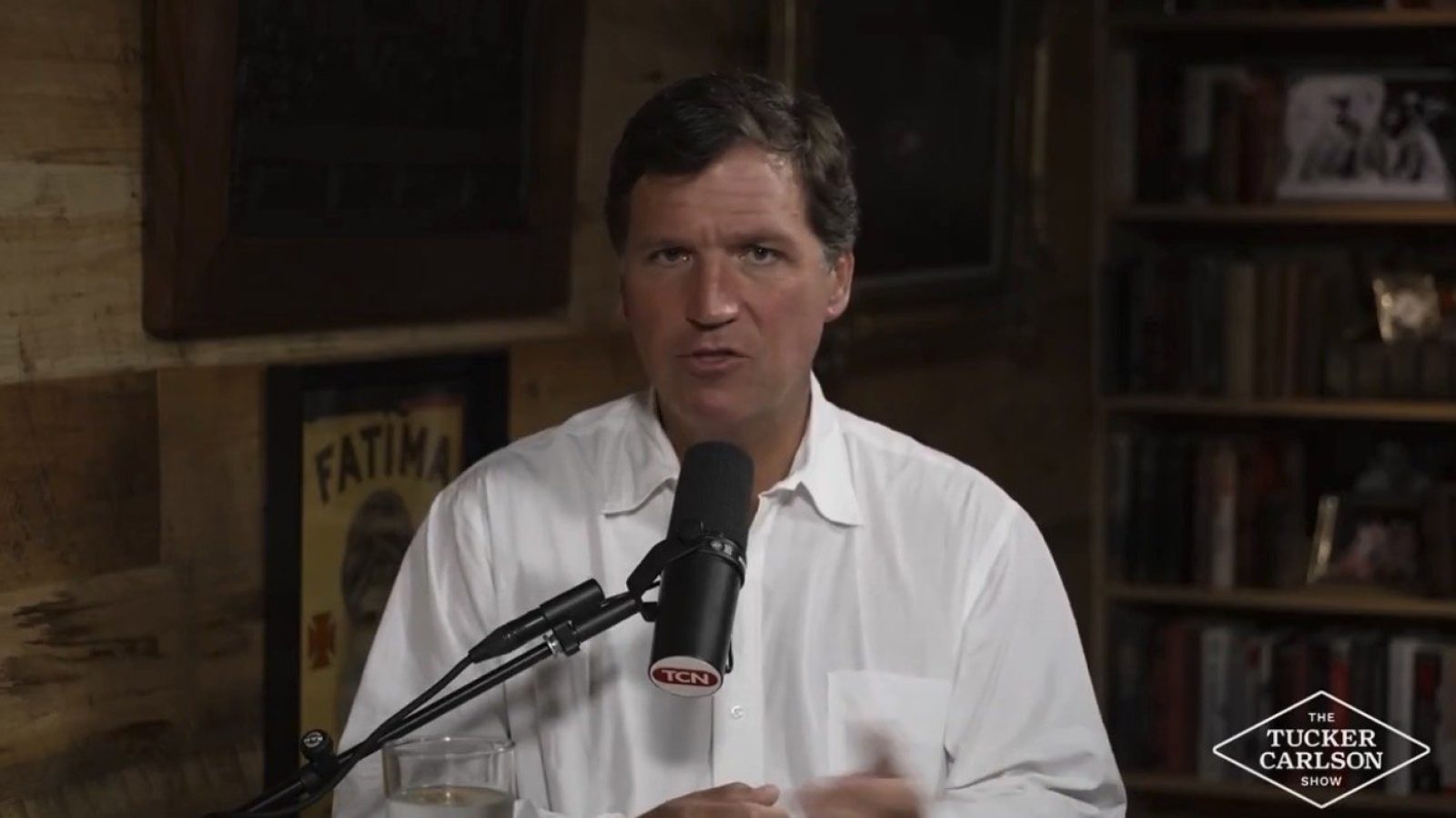 Tucker Carlson discussing current events on his show, seated at a desk with a microphone and a glass, in a cozy, wood-paneled setting.