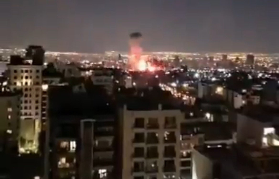 Nighttime cityscape showing a fire or explosion with bright flames illuminating the skyline, surrounded by urban buildings and distant city lights.