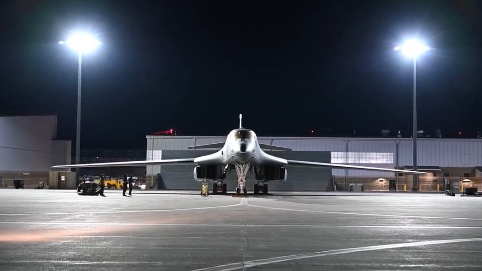 A military aircraft on a tarmac at night, with personnel and equipment visible under bright lights.