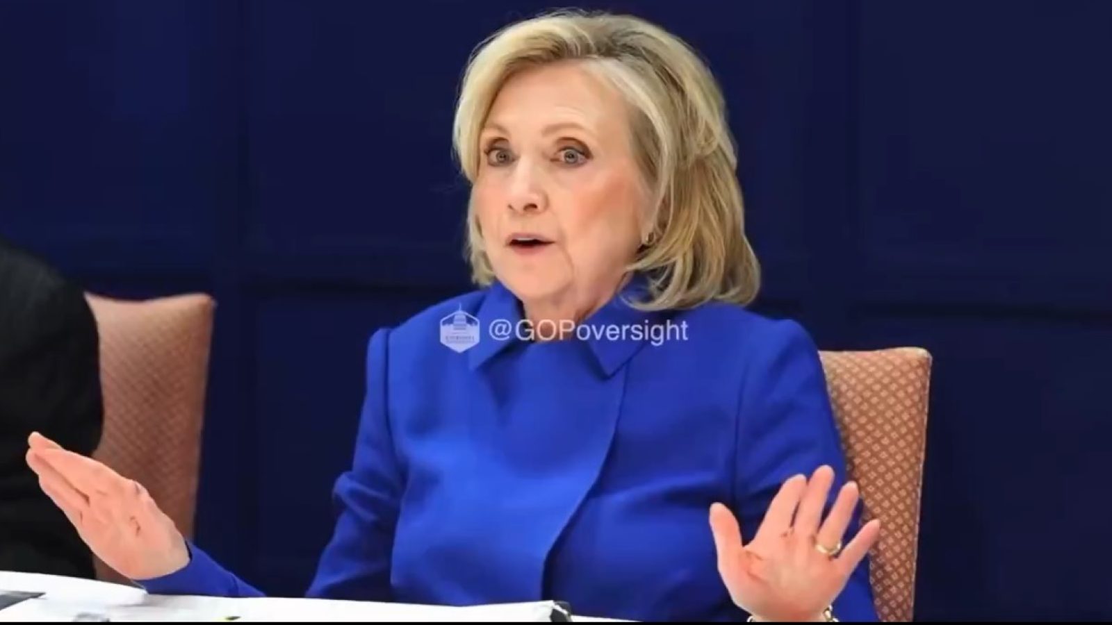 Hillary Clinton speaking during a discussion, wearing a blue blazer and gesturing with her hands, emphasizing her points.
