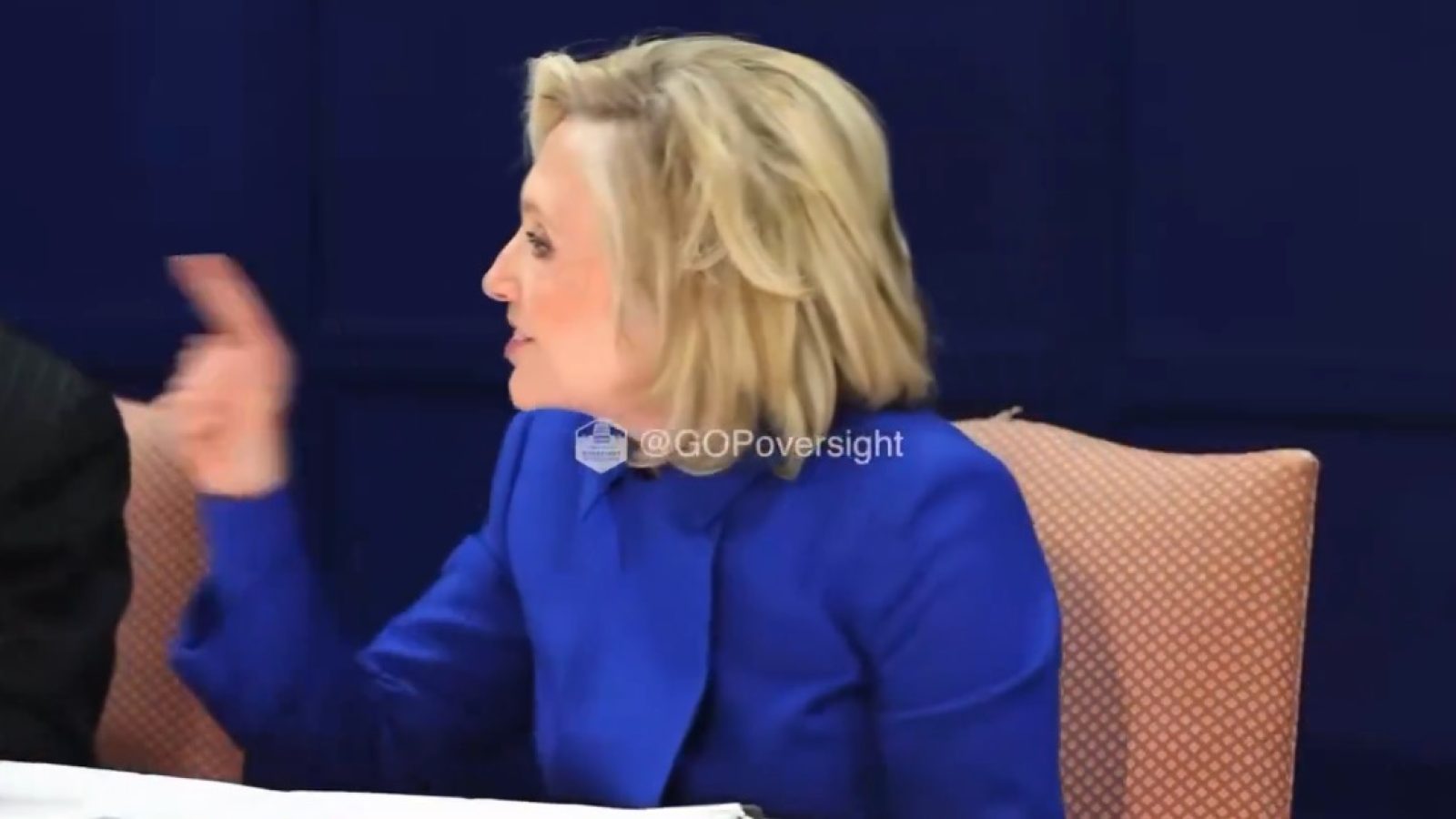 Hillary Clinton gestures while speaking at a meeting, wearing a blue blazer, with a focused expression in a professional setting.