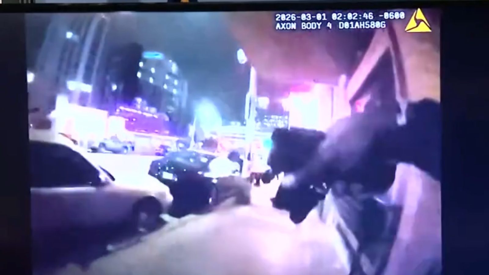 Body camera footage capturing a nighttime police encounter on a city street, featuring illuminated signs and vehicles in the background.