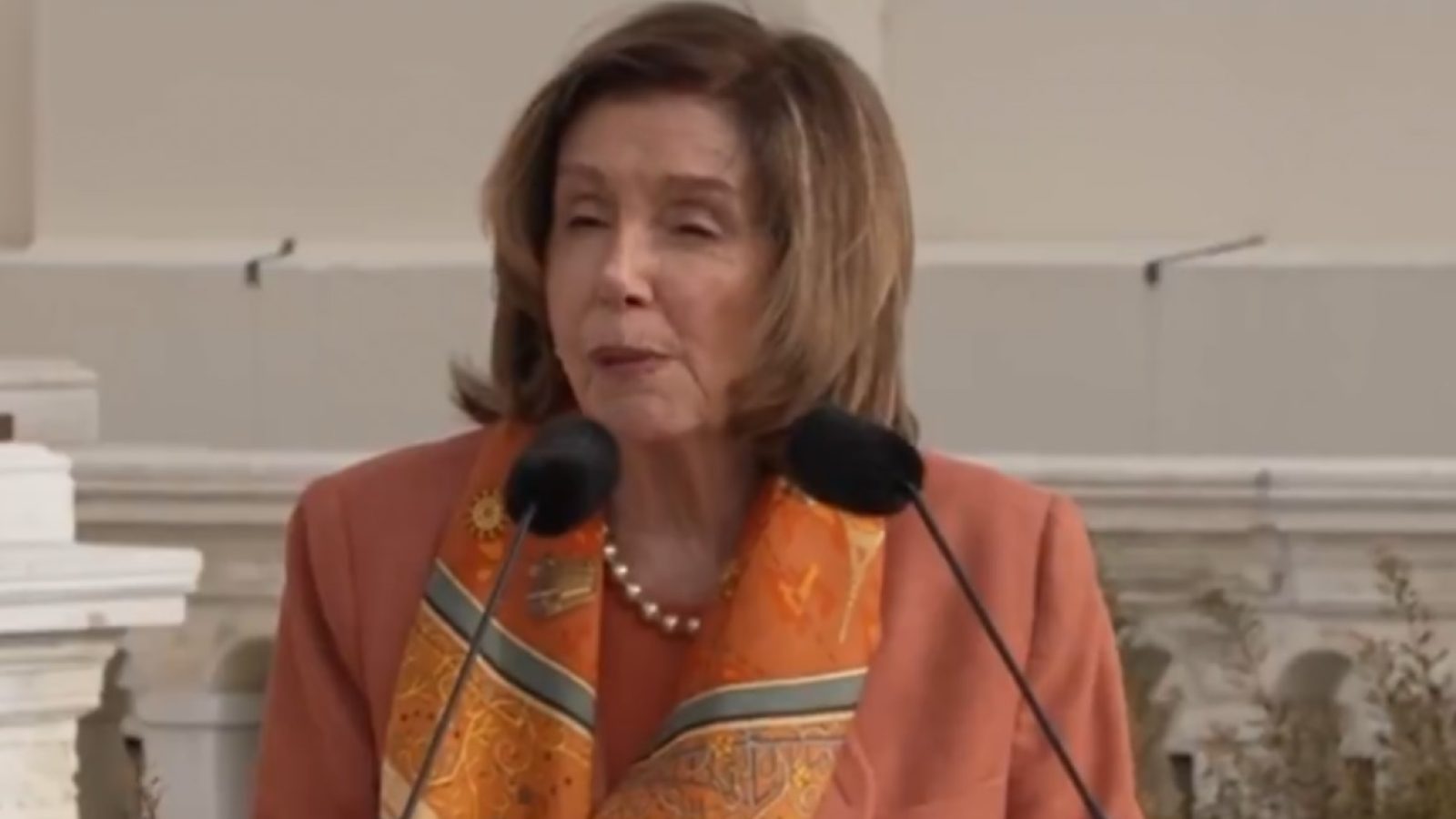 Speaker delivering a speech outdoors, wearing an orange blazer and a decorative scarf, with microphones positioned in front of her.