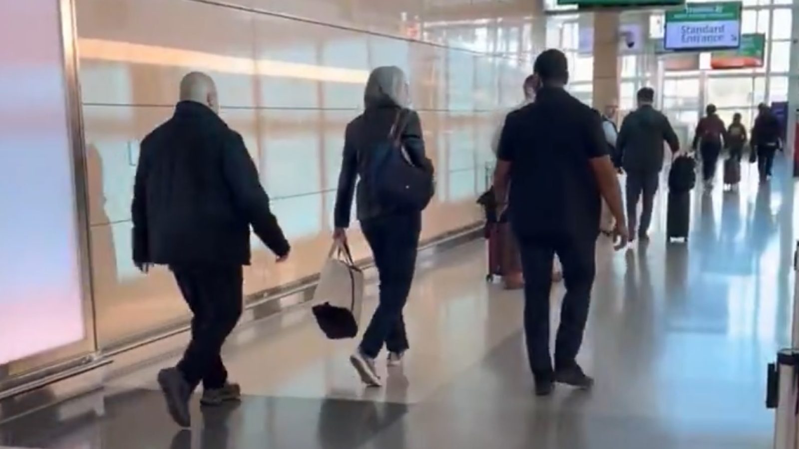 People walking through a busy airport terminal, with luggage and signs in the background.
