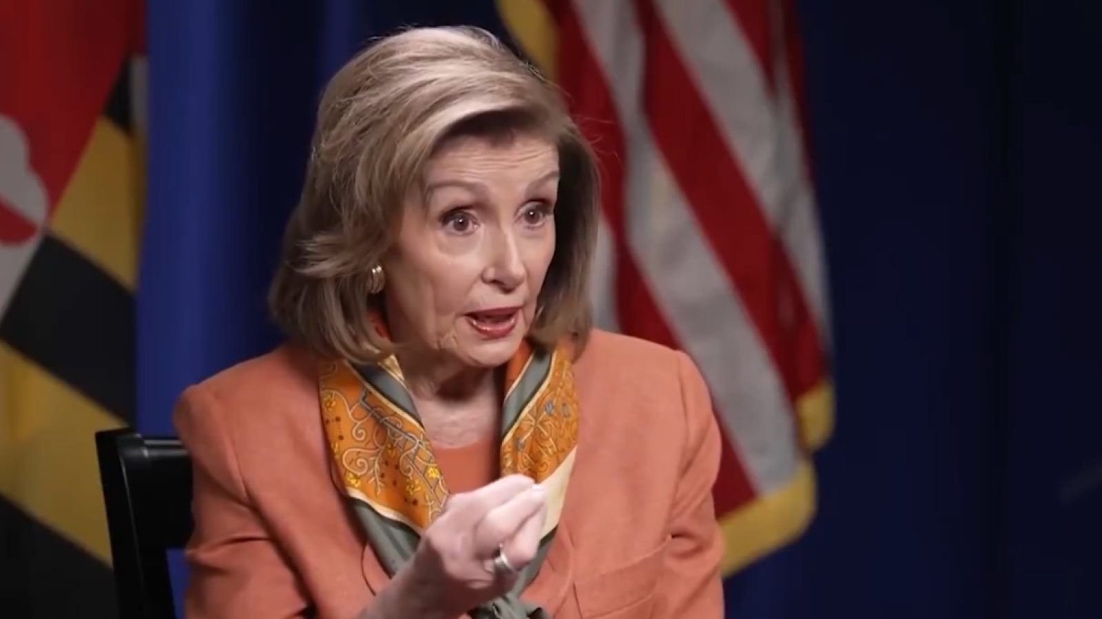 Woman with blonde hair wearing an orange blazer and colorful scarf gestures while speaking, with flags in the background, in a discussion setting.