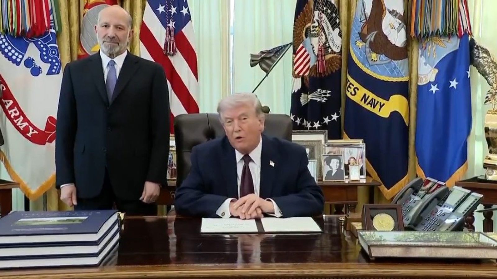 Donald Trump seated at a desk in the Oval Office, signing documents with an official standing beside him, surrounded by military flags and personal items.