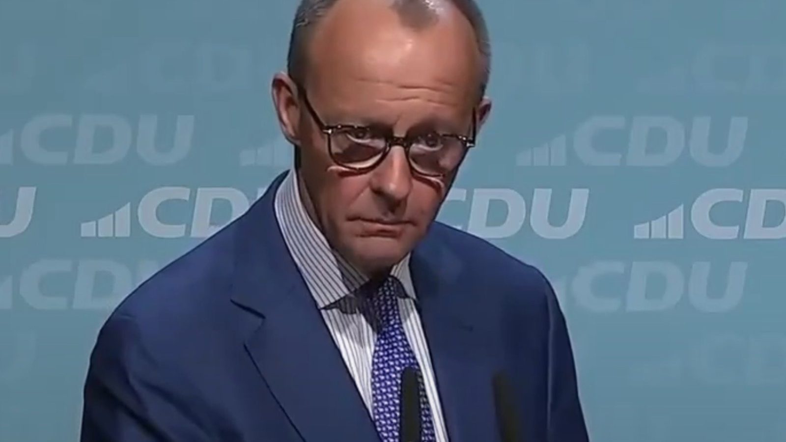 Politician in a blue suit and glasses stands at a podium during a CDU press conference, presenting a serious expression against a light blue background.